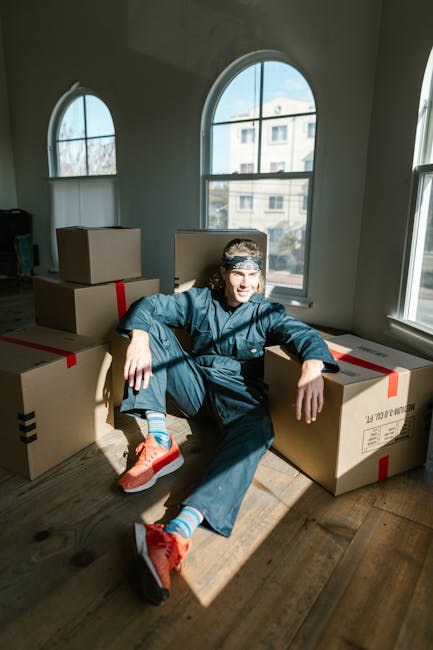 A young man with a beard and curly hair, dressed in a dark blue t-shirt and matching trousers, is seated on a wooden stool inside a room with a textured light blue wall. He is holding a clipboard and 