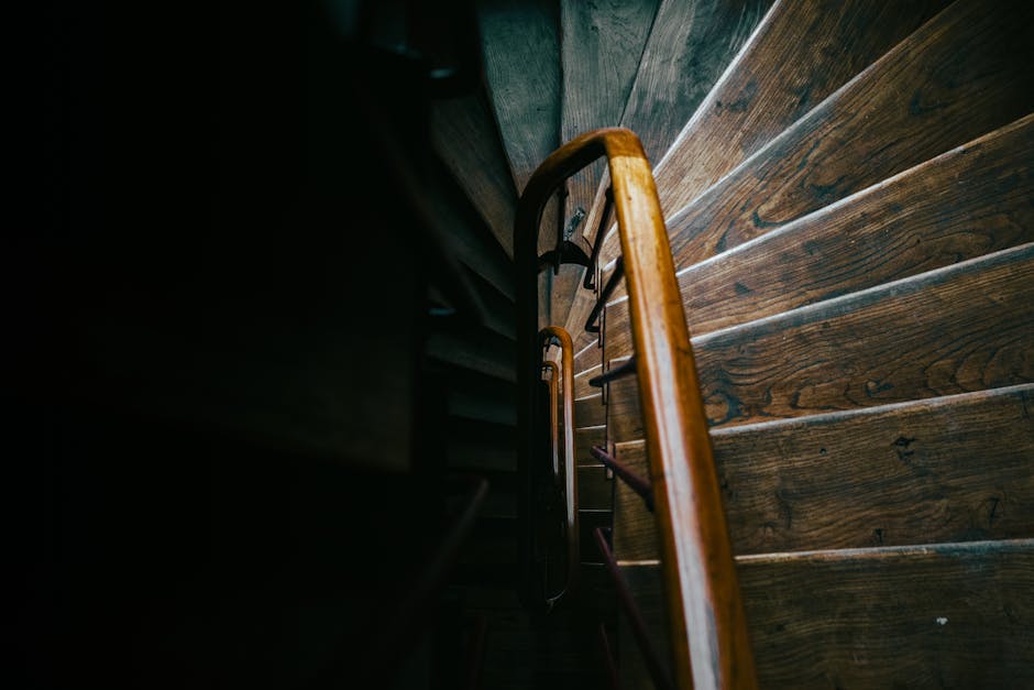 Interior view of a residential staircase with concrete steps leading upwards, located next to a large window with a black metal railing running alongside the stairs. The window allows natural light to
