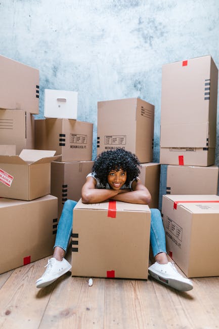 A young woman with curly black hair, wearing a white t-shirt, blue jeans, and white sneakers, is sitting on the wooden floor of a room surrounded by numerous large cardboard boxes. The boxes vary in s
