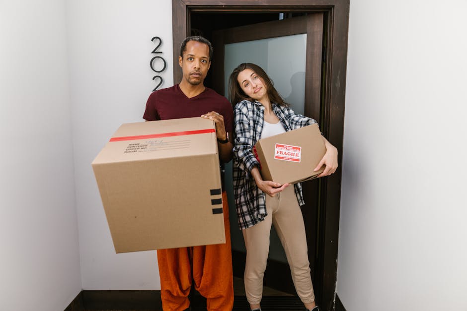 A young woman with curly black hair, wearing a white t-shirt, blue jeans, and white sneakers, is sitting on the wooden floor of a room surrounded by numerous large cardboard boxes. The boxes vary in s