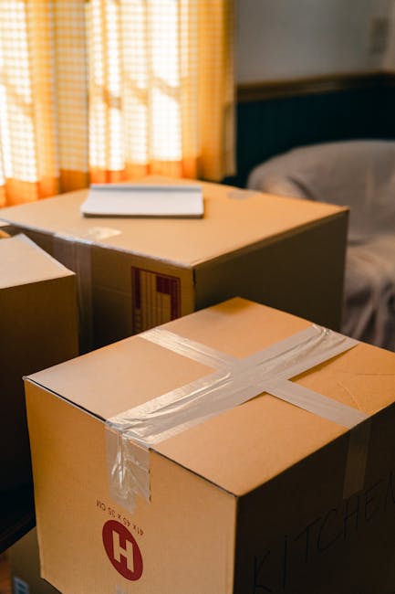 A collection of unpacked cardboard moving boxes of various sizes positioned on the floor and near a windowsill in a residential room. The boxes are partially open, revealing packing materials such as